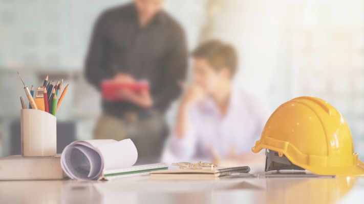 Schreibtisch mit Bauhelm, technischen Zeichnungen und Bürobedarf – im Hintergrund zwei Personen im Gespräch – Symbolbild für die Tätigkeit als Projektleiter TGA im Bereich Gebäudetechnik